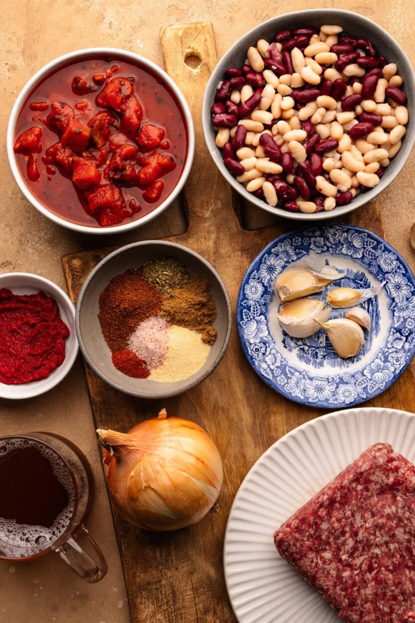 Overhead shot of all the beef chili ingredients laid out on a wooden cutting board — ground beef, canned tomatoes, kidney beans, black beans, onion, garlic, bell pepper, and small bowls of chili spices