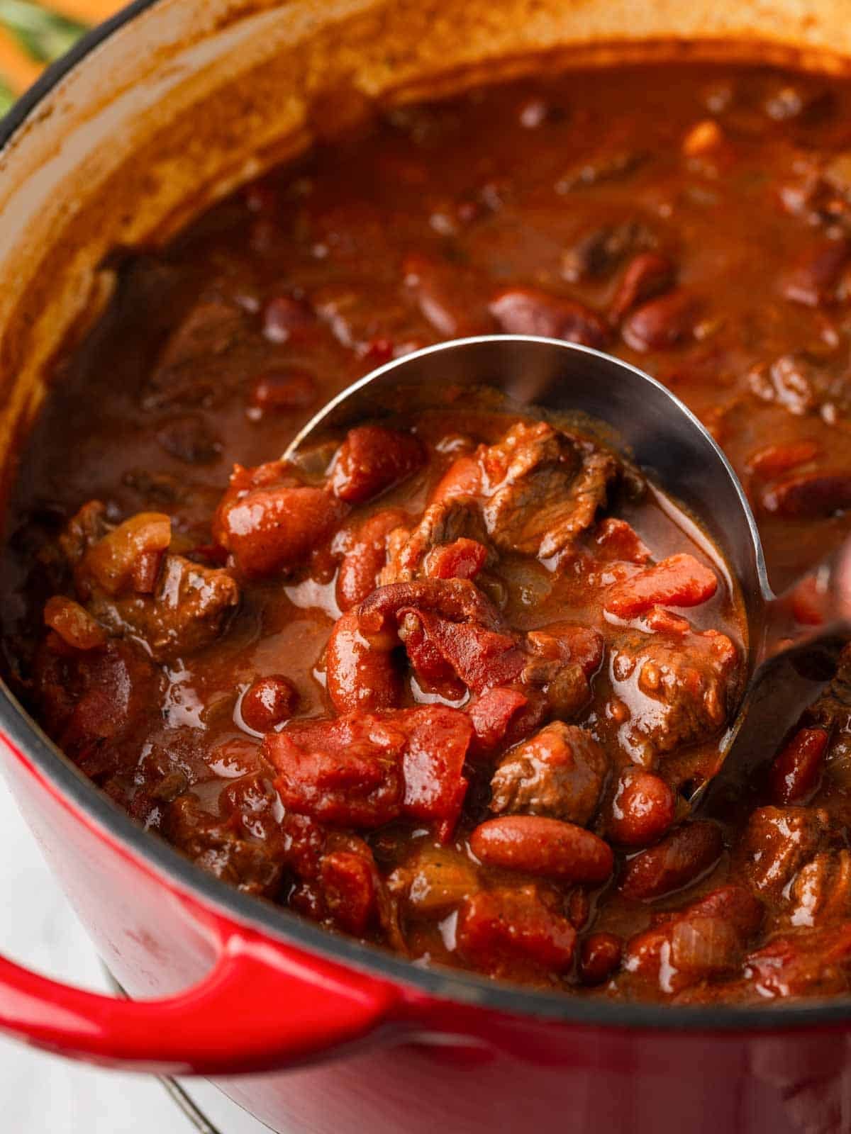 A close-up shot of a ladle scooping thick beef chili from a Dutch oven, showing the rich sauce, ground beef, and whole beans in vivid detail