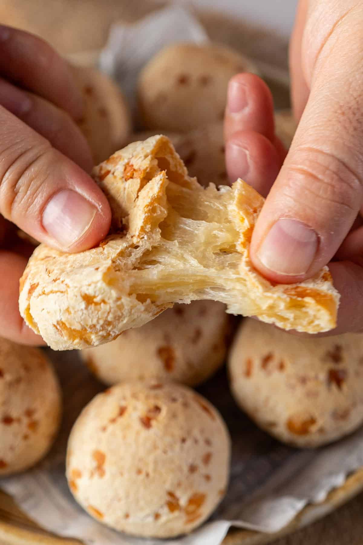 Close-up of a single Brazilian Pão de Queijo roll torn open in half, showing the golden crispy exterior and the stretchy, gooey cheesy interior, served on a dark wooden board