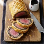 Sliced British Beef Wellington on a wooden board, showing a perfect medium-rare pink center wrapped in golden puff pastry and mushroom duxelles, garnished with fresh thyme