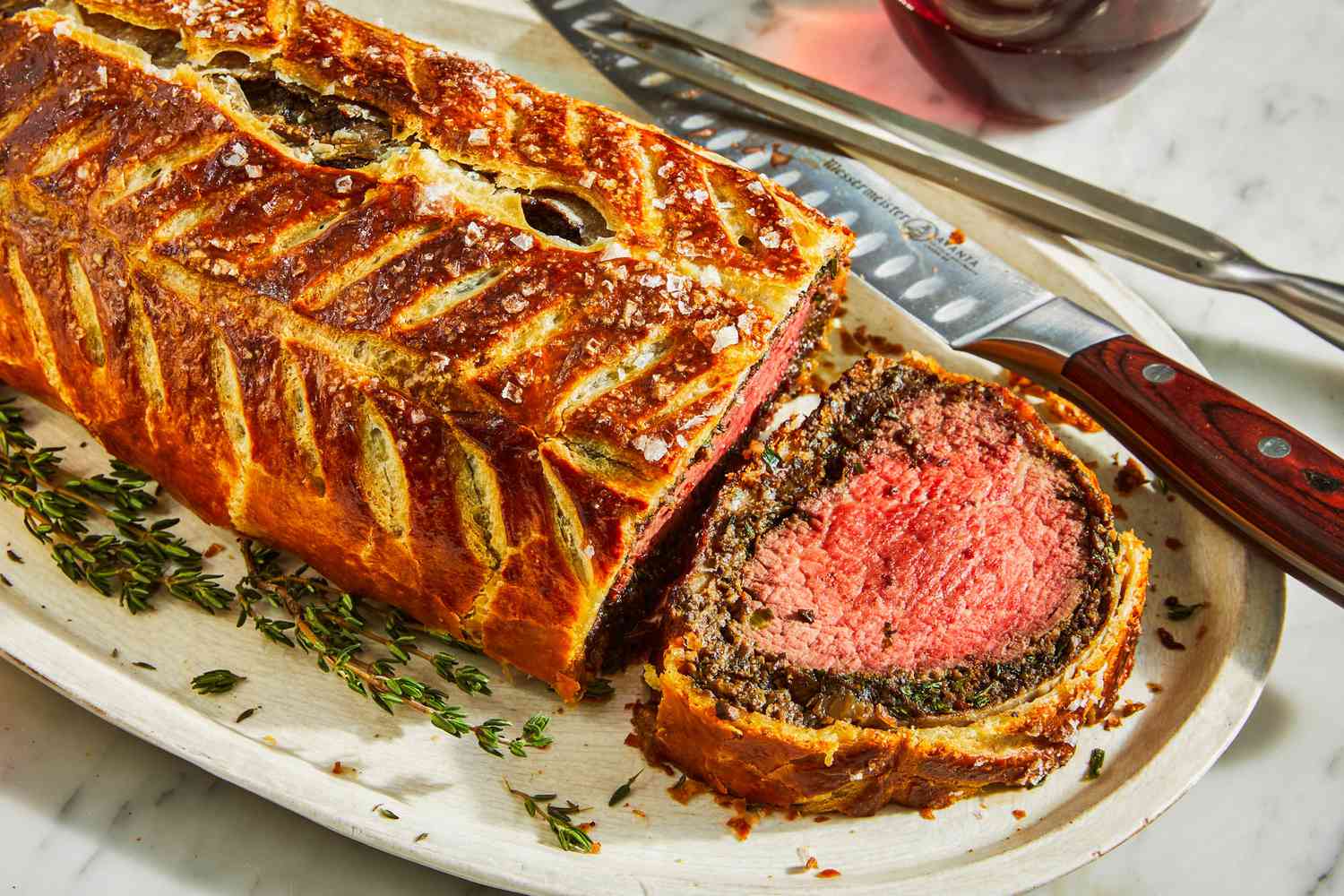 Step-by-step flat lay of British Beef Wellington assembly — mushroom duxelles spread over prosciutto slices on plastic wrap, with seared beef tenderloin ready to be rolled, alongside puff pastry sheet and egg wash