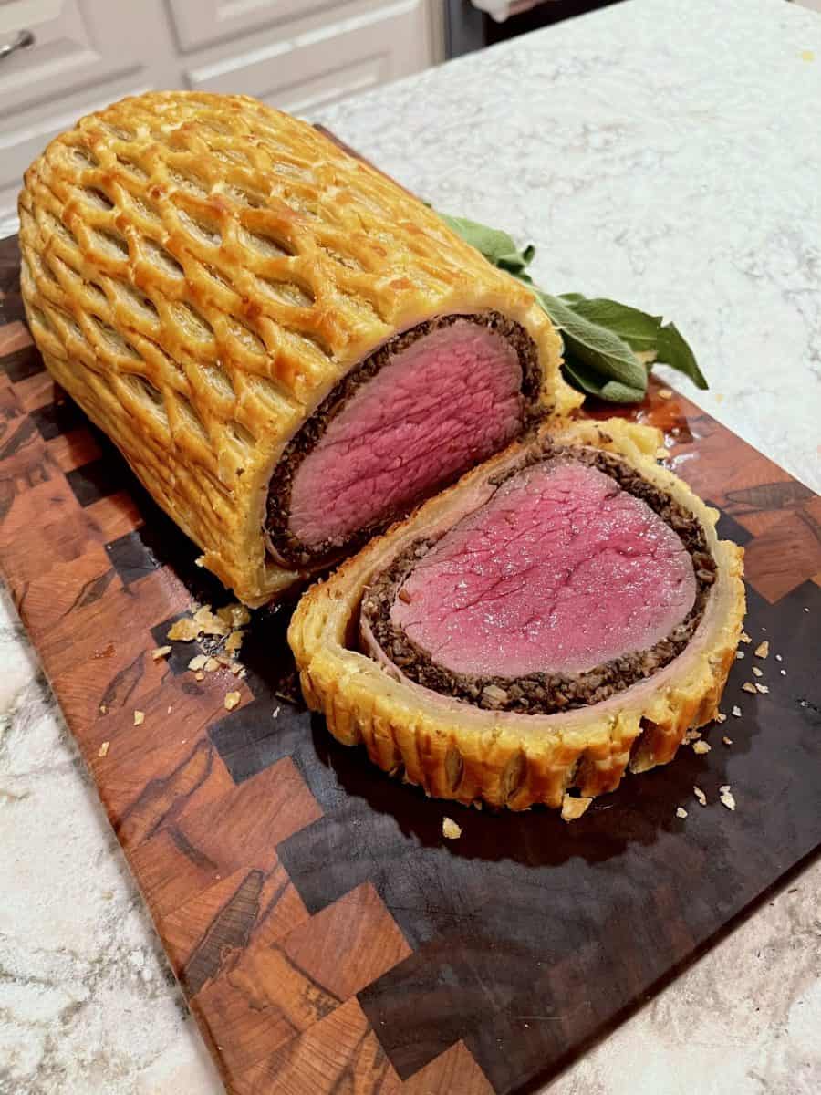 Close-up cross-section of British Beef Wellington showing a vivid medium-rare pink tenderloin center, dark mushroom duxelles layer, prosciutto wrapping, and shatteringly crisp golden puff pastry exterior, plated with red wine jus