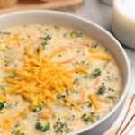 A bowl of rich, golden broccoli cheddar soup topped with shredded cheddar and freshly cracked black pepper, served in a white ceramic bowl with a crusty bread roll on the side