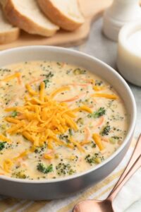 A bowl of rich, golden broccoli cheddar soup topped with shredded cheddar and freshly cracked black pepper, served in a white ceramic bowl with a crusty bread roll on the side