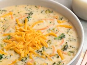 A bowl of rich, golden broccoli cheddar soup topped with shredded cheddar and freshly cracked black pepper, served in a white ceramic bowl with a crusty bread roll on the side