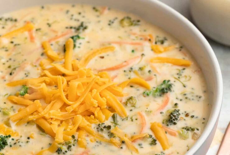 A bowl of rich, golden broccoli cheddar soup topped with shredded cheddar and freshly cracked black pepper, served in a white ceramic bowl with a crusty bread roll on the side