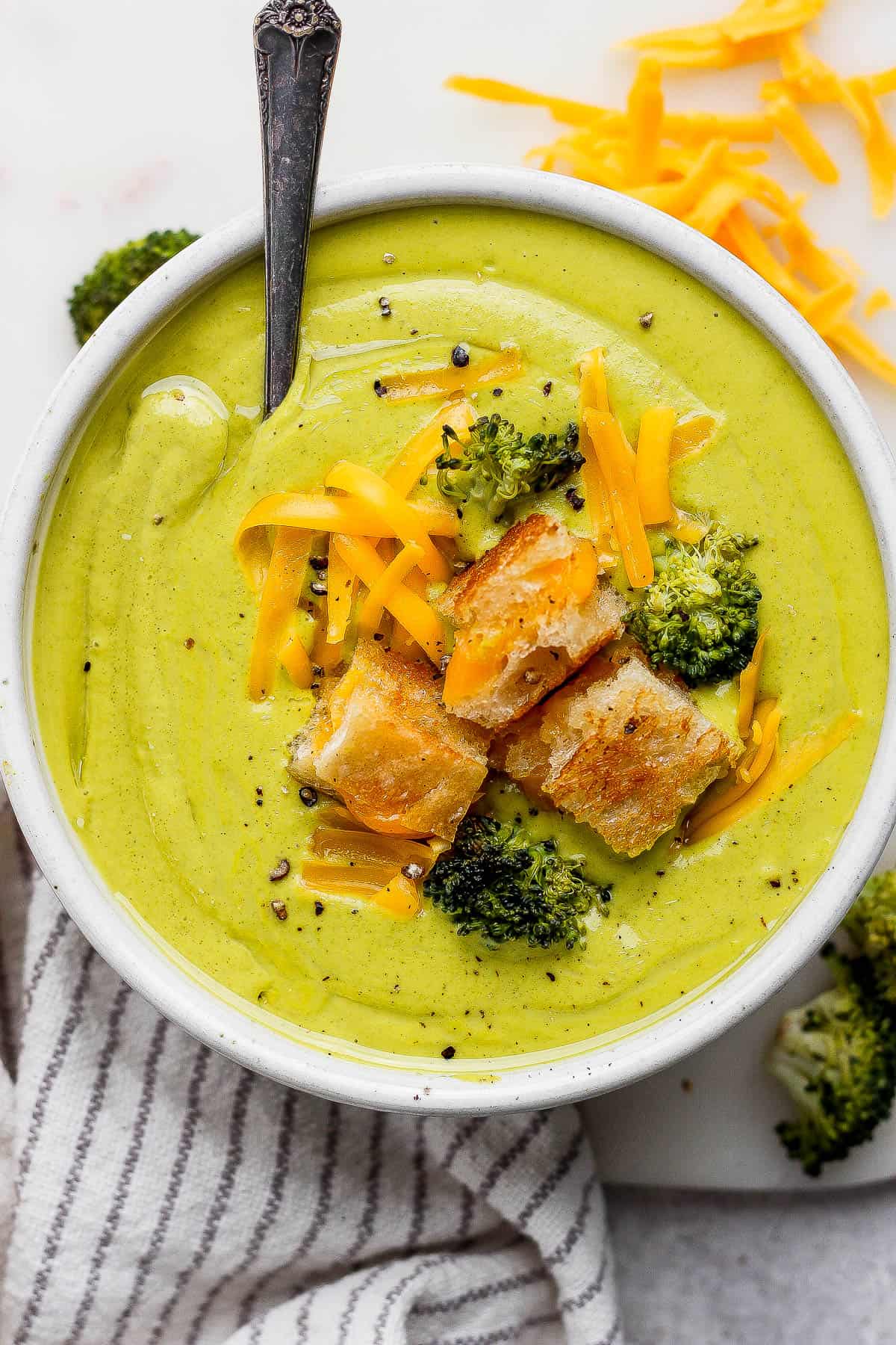 An overhead flat-lay of broccoli cheddar soup ingredients: a block of sharp cheddar, fresh broccoli florets, diced yellow onion, garlic cloves, heavy cream, butter, and chicken broth arranged on a wooden cutting board