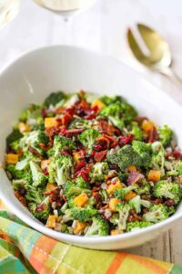 Overhead shot of broccoli salad with bacon, cheddar, and sunflower seeds in a large white serving bowl, garnished with extra bacon crumbles, styled on a rustic wooden picnic table