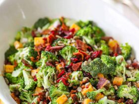 Overhead shot of broccoli salad with bacon, cheddar, and sunflower seeds in a large white serving bowl, garnished with extra bacon crumbles, styled on a rustic wooden picnic table