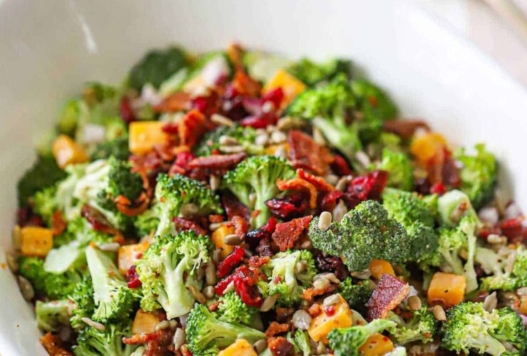 Overhead shot of broccoli salad with bacon, cheddar, and sunflower seeds in a large white serving bowl, garnished with extra bacon crumbles, styled on a rustic wooden picnic table