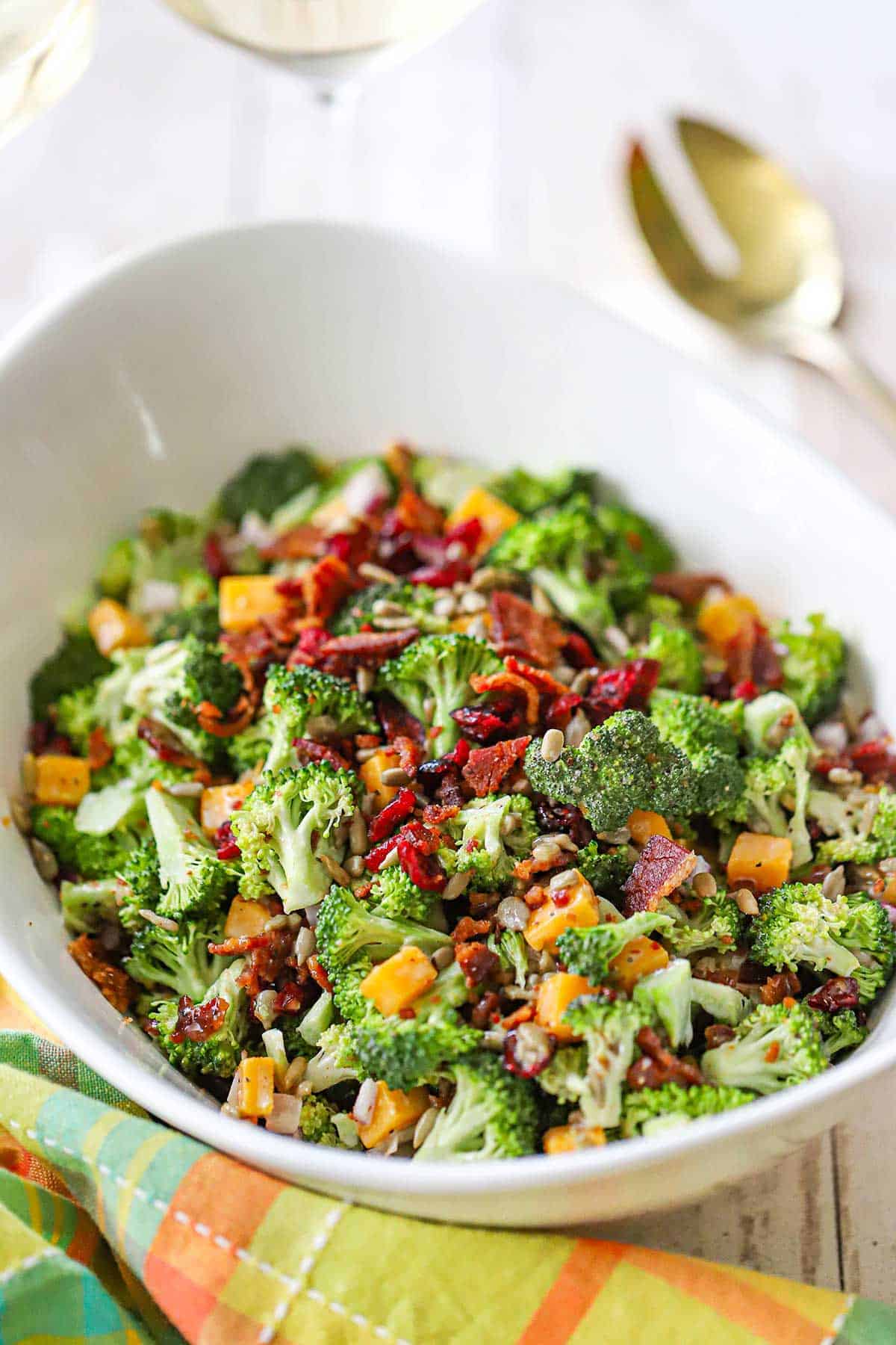 Overhead shot of broccoli salad with bacon, cheddar, and sunflower seeds in a large white serving bowl, garnished with extra bacon crumbles, styled on a rustic wooden picnic table