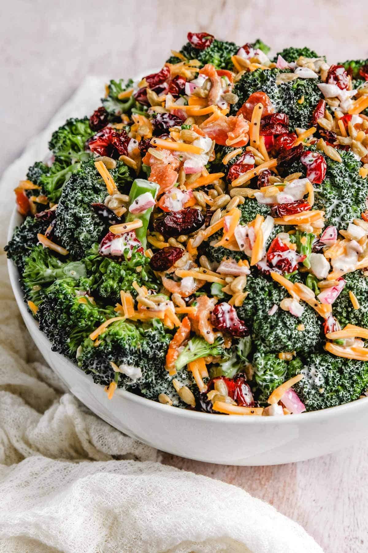 Close-up shot of a large serving spoon scooping broccoli salad from a white bowl, showing creamy dressing coating the florets, visible bacon crumbles, cheddar cubes, and sunflower seeds