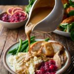 Brown butter turkey gravy being poured from a white gravy boat over sliced roasted turkey and mashed potatoes on a rustic Thanksgiving table
