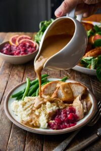 Brown butter turkey gravy being poured from a white gravy boat over sliced roasted turkey and mashed potatoes on a rustic Thanksgiving table