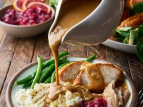 Brown butter turkey gravy being poured from a white gravy boat over sliced roasted turkey and mashed potatoes on a rustic Thanksgiving table