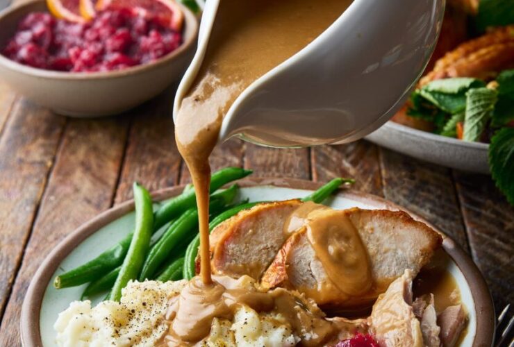 Brown butter turkey gravy being poured from a white gravy boat over sliced roasted turkey and mashed potatoes on a rustic Thanksgiving table