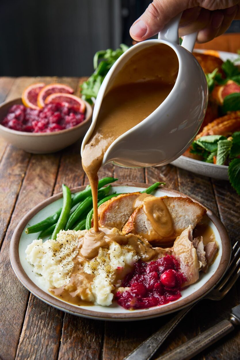 Brown butter turkey gravy being poured from a white gravy boat over sliced roasted turkey and mashed potatoes on a rustic Thanksgiving table