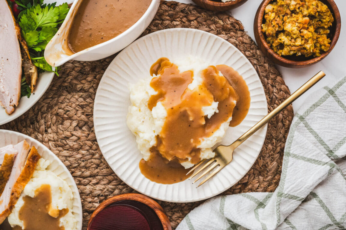 Overhead flat-lay of brown butter turkey gravy ingredients including a stick of butter browning in a saucepan, turkey drippings in a measuring cup, fresh thyme, flour, and stock on a wooden kitchen surface