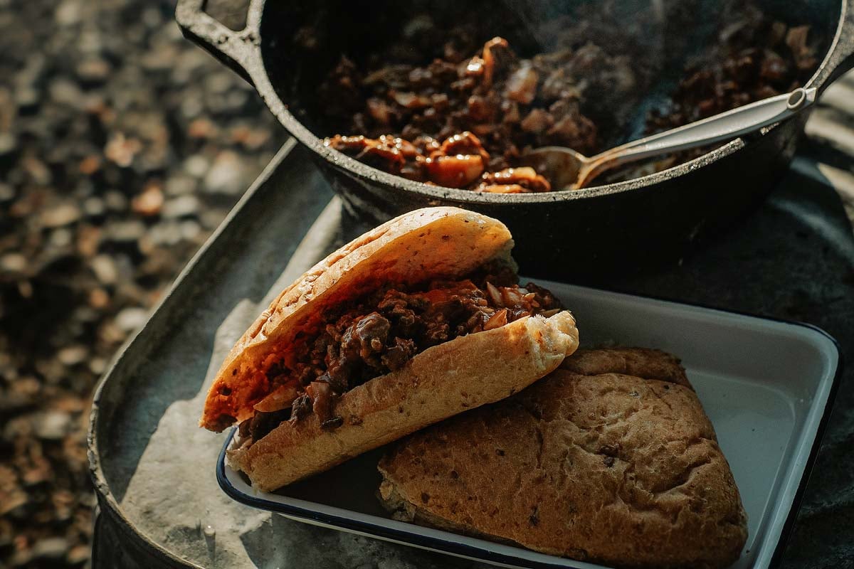 Dutch oven filled with simmering tangy ground beef sloppy joe filling, surrounded by campfire ingredients including onions, paprika, Worcestershire, and ketchup