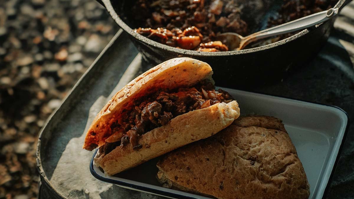 Close-up of a Campfire Sloppy Joe on a toasted brioche bun with melted cheddar and pickles, held over a glowing campfire in the background