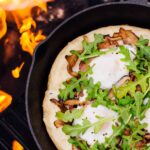 Cast iron skillet pizza with golden crispy crust, bubbling mozzarella cheese, and fresh basil leaves served on a rustic wooden table at a campsite with firelight in the background