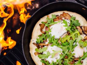 Cast iron skillet pizza with golden crispy crust, bubbling mozzarella cheese, and fresh basil leaves served on a rustic wooden table at a campsite with firelight in the background