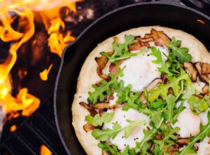 Cast iron skillet pizza with golden crispy crust, bubbling mozzarella cheese, and fresh basil leaves served on a rustic wooden table at a campsite with firelight in the background