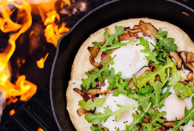 Cast iron skillet pizza with golden crispy crust, bubbling mozzarella cheese, and fresh basil leaves served on a rustic wooden table at a campsite with firelight in the background