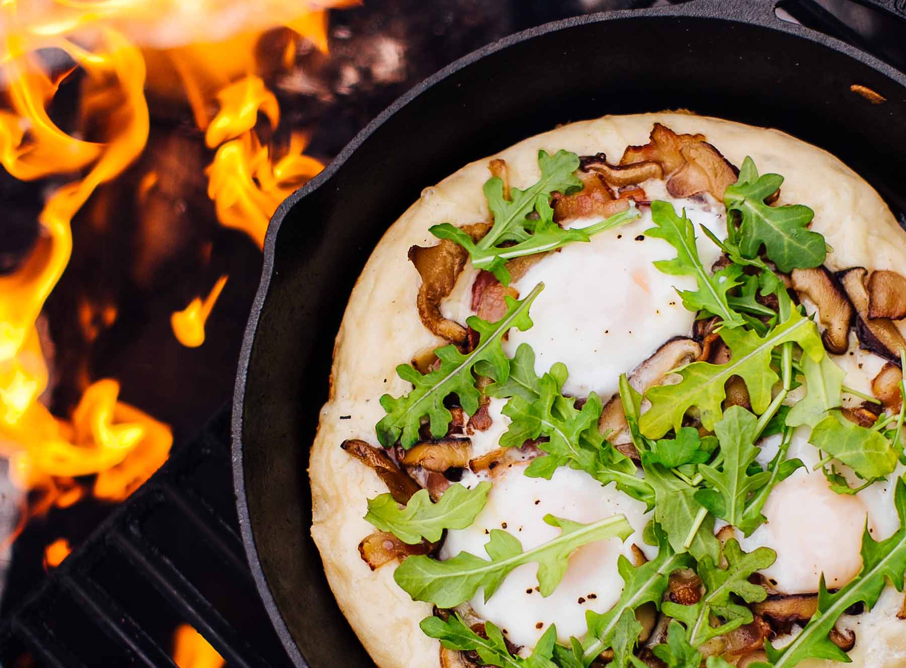 Cast iron skillet pizza with golden crispy crust, bubbling mozzarella cheese, and fresh basil leaves served on a rustic wooden table at a campsite with firelight in the background