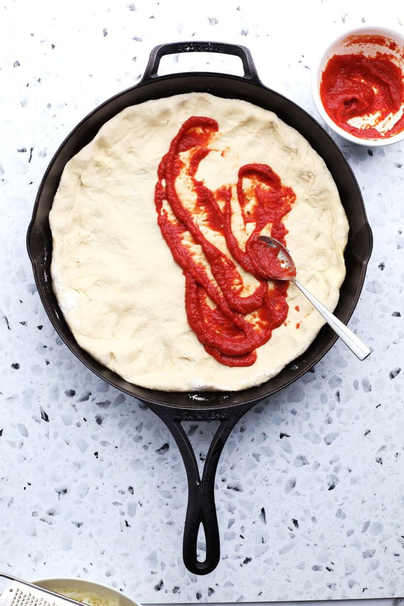 Overhead flat lay of pre-made pizza dough ball, jar of marinara sauce, shredded mozzarella, pepperoni slices, olive oil bottle, and dried herbs arranged on a rustic wooden surface with a cast iron skillet beside them