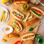 Chicago Style Hot Dogs beautifully plated on a wooden board with all seven toppings visible — yellow mustard, neon green relish, diced onion, tomato wedges, dill pickle spear, sport peppers, and celery salt on a poppy seed bun