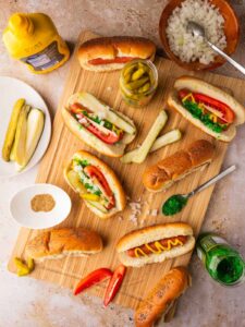 Chicago Style Hot Dogs beautifully plated on a wooden board with all seven toppings visible — yellow mustard, neon green relish, diced onion, tomato wedges, dill pickle spear, sport peppers, and celery salt on a poppy seed bun