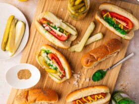 Chicago Style Hot Dogs beautifully plated on a wooden board with all seven toppings visible — yellow mustard, neon green relish, diced onion, tomato wedges, dill pickle spear, sport peppers, and celery salt on a poppy seed bun