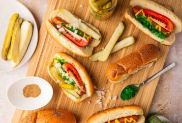 Chicago Style Hot Dogs beautifully plated on a wooden board with all seven toppings visible — yellow mustard, neon green relish, diced onion, tomato wedges, dill pickle spear, sport peppers, and celery salt on a poppy seed bun
