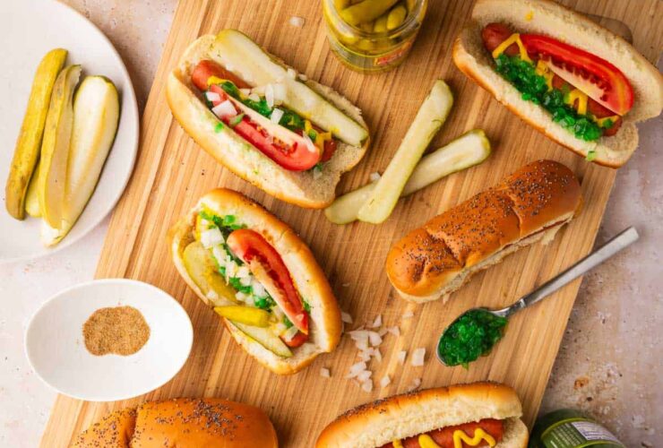 Chicago Style Hot Dogs beautifully plated on a wooden board with all seven toppings visible — yellow mustard, neon green relish, diced onion, tomato wedges, dill pickle spear, sport peppers, and celery salt on a poppy seed bun