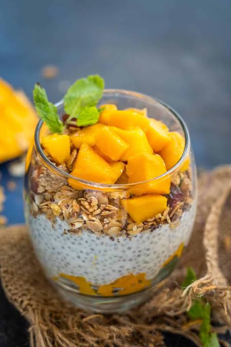Overhead shot of ingredients for coconut chia pudding parfait including canned coconut milk, chia seeds, fresh mango slices, granola, and toasted coconut flakes arranged on a white marble surface