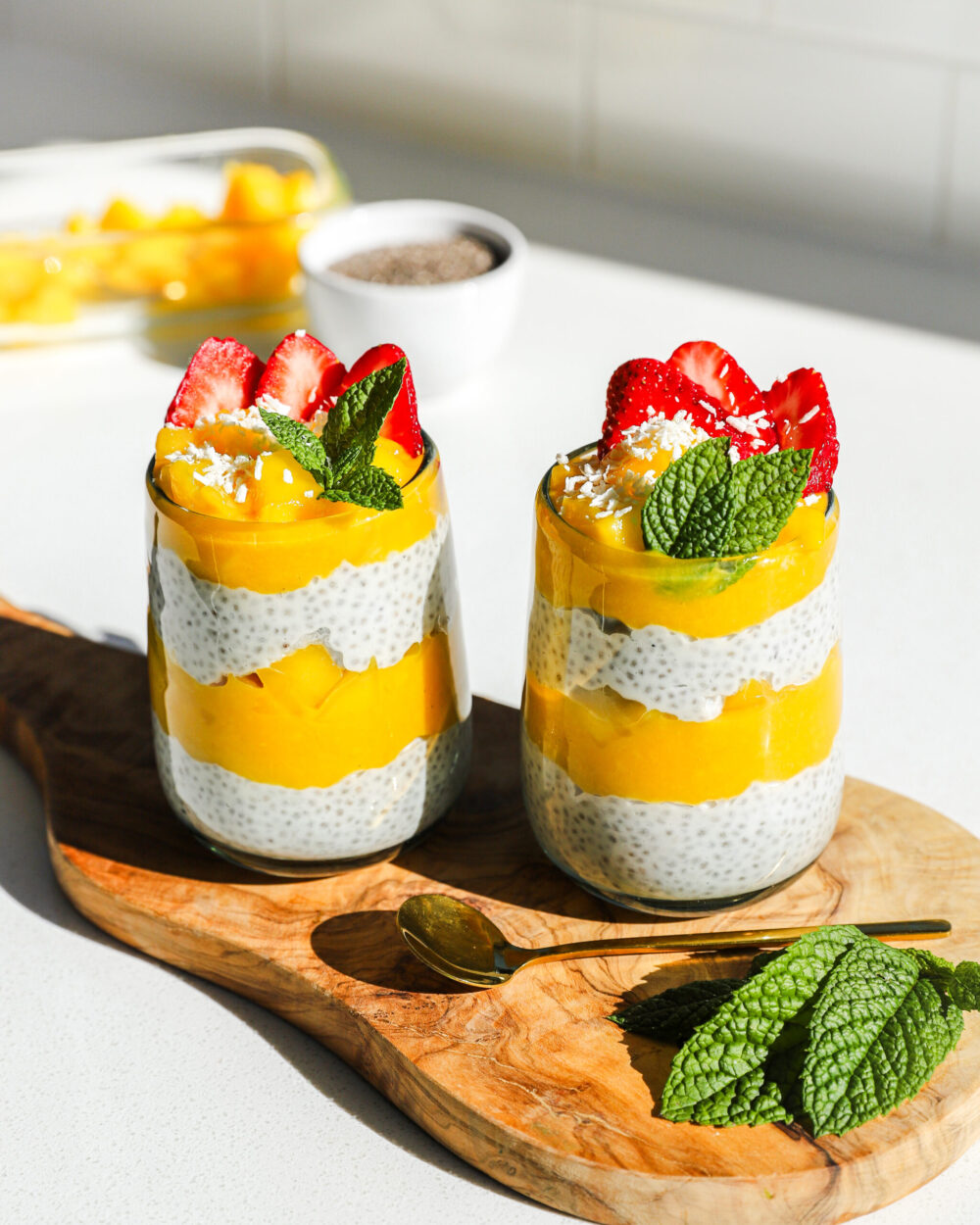 Close-up of a single coconut chia pudding parfait in a clear glass jar showing distinct layers of creamy white chia pudding, golden mango chunks, crunchy granola, and toasted coconut flakes with a passionfruit drizzle on top