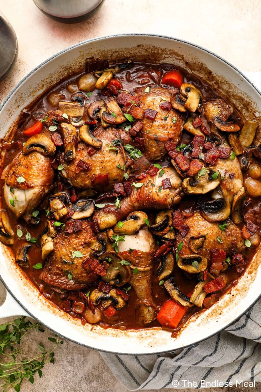Step-by-step process shot showing chicken pieces being seared golden brown in a Dutch oven with rendered bacon lardons and pearl onions