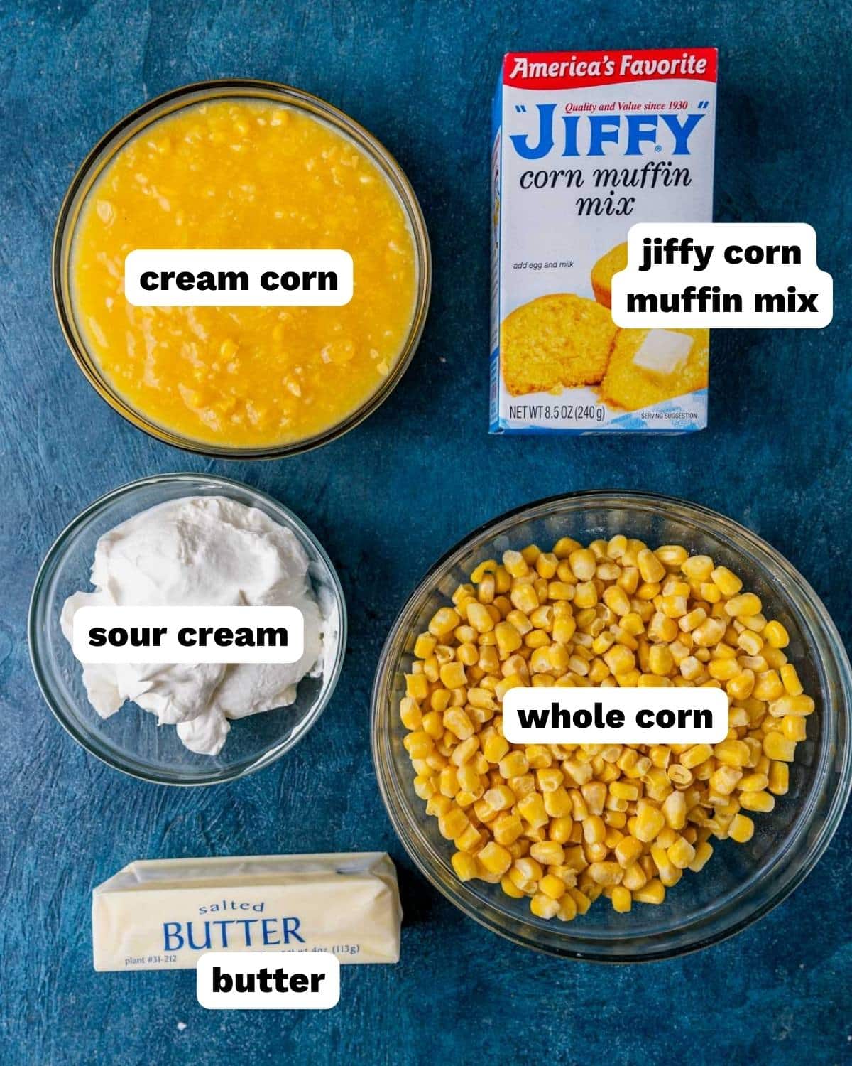Overhead flat-lay of corn casserole ingredients: two cans of corn, sour cream, eggs, melted butter, Jiffy mix, and a glass baking dish on a linen surface