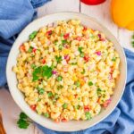 A large white bowl of creamy macaroni salad with colorful vegetables, garnished with paprika and fresh parsley, shot overhead on a rustic wooden picnic table
