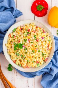 A large white bowl of creamy macaroni salad with colorful vegetables, garnished with paprika and fresh parsley, shot overhead on a rustic wooden picnic table