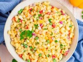 A large white bowl of creamy macaroni salad with colorful vegetables, garnished with paprika and fresh parsley, shot overhead on a rustic wooden picnic table