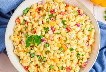 A large white bowl of creamy macaroni salad with colorful vegetables, garnished with paprika and fresh parsley, shot overhead on a rustic wooden picnic table