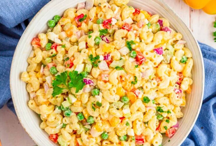 A large white bowl of creamy macaroni salad with colorful vegetables, garnished with paprika and fresh parsley, shot overhead on a rustic wooden picnic table