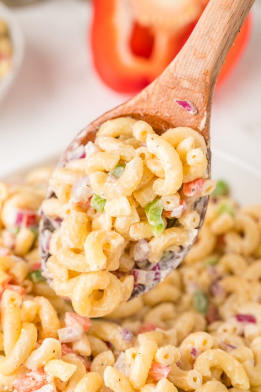 A close-up spoonful of creamy macaroni salad being lifted from the bowl, showing the glossy mayo dressing coating each elbow noodle, with flecks of red bell pepper and celery visible