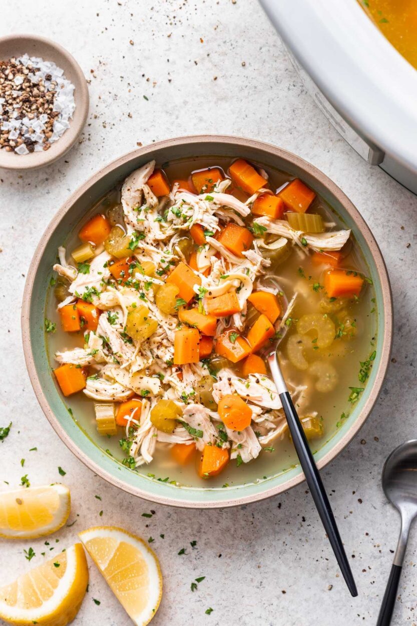 A close-up of a ladle lifting a generous scoop of Crockpot Chicken Noodle Soup from a white ceramic crockpot, showing golden broth, shredded chicken, carrots, celery, and egg noodles