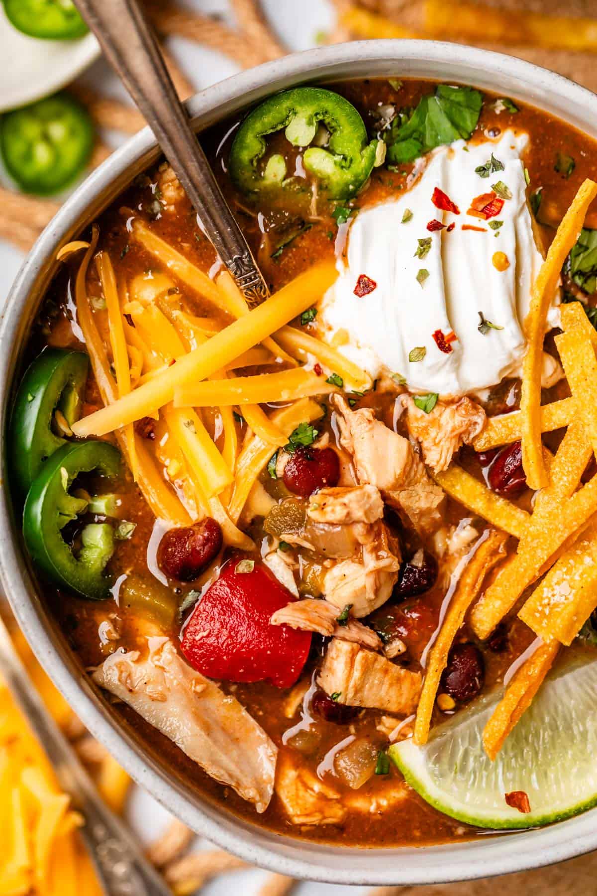 Close-up of a bowl of Crockpot Chicken Tortilla Soup with shredded chicken visible in a rich, smoky broth, topped with a dollop of sour cream, crispy golden tortilla strips, shredded orange cheese, and a lime wedge on the side
