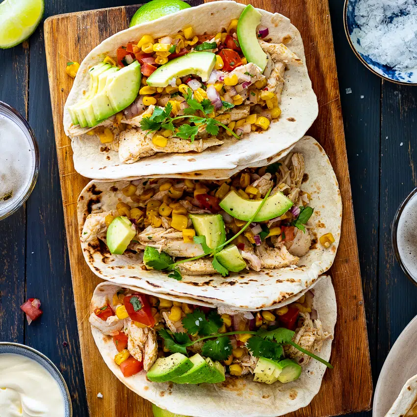 Close-up of pulled salsa chicken in warm corn tortillas topped with diced avocado, fresh cilantro, and a drizzle of sour cream, shot against a dark moody background