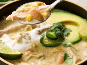 Crockpot white chicken chili served in a rustic bowl topped with sour cream, shredded cheese, jalapeño slices, cilantro, and a squeeze of lime on a dark moody surface