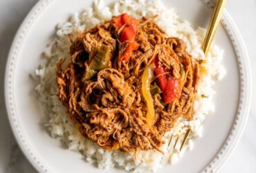 Cuban Ropa Vieja served over white rice in a rustic bowl, garnished with green olives and fresh cilantro, rich red tomato sauce glistening under warm light
